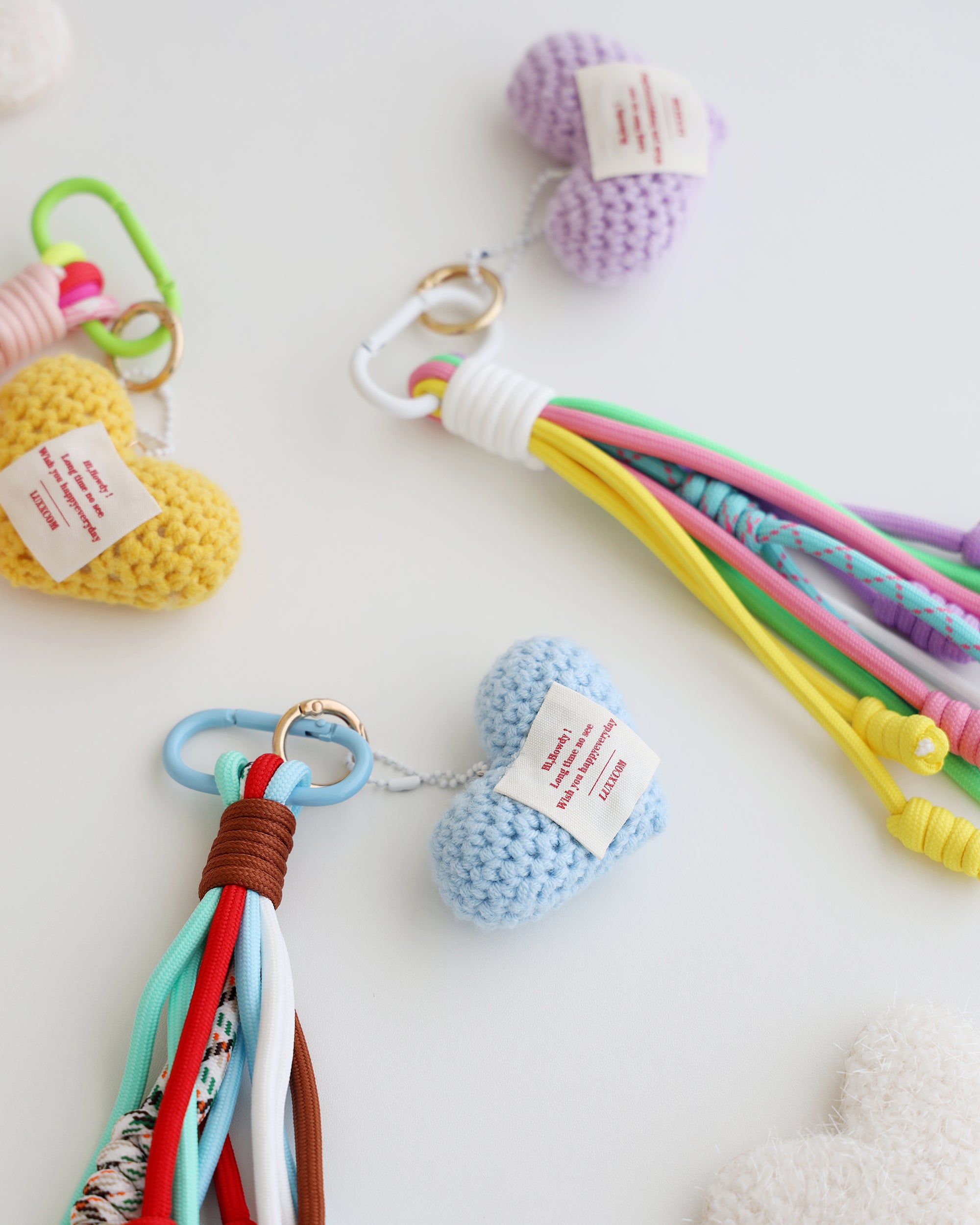 Close-up of three keychain charms made with multiple knotted cord tassels and a crocheted heart. Each charm includes a metal carabiner clip for attaching to bags, keys, or other items. The tassels are made of multi-color cords, and the crochet heart features a stitched label.