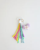 A keychain charm made with multiple knotted cord tassels and a lavender crocheted heart which includes a white  metal carabiner clip for attaching to bags, keys, or other items. The tassels are made of multi-color cords, and the crochet heart features a stitched label.
