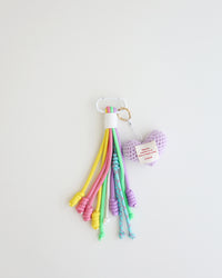 A keychain charm made with multiple knotted cord tassels and a lavender crocheted heart which includes a white  metal carabiner clip for attaching to bags, keys, or other items. The tassels are made of multi-color cords, and the crochet heart features a stitched label.