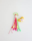 A keychain charm made with multiple knotted cord tassels and a soft yellow crocheted heart which includes a pastel green metal carabiner clip for attaching to bags, keys, or other items. The tassels are made of multi-color cords, and the crochet heart features a stitched label.