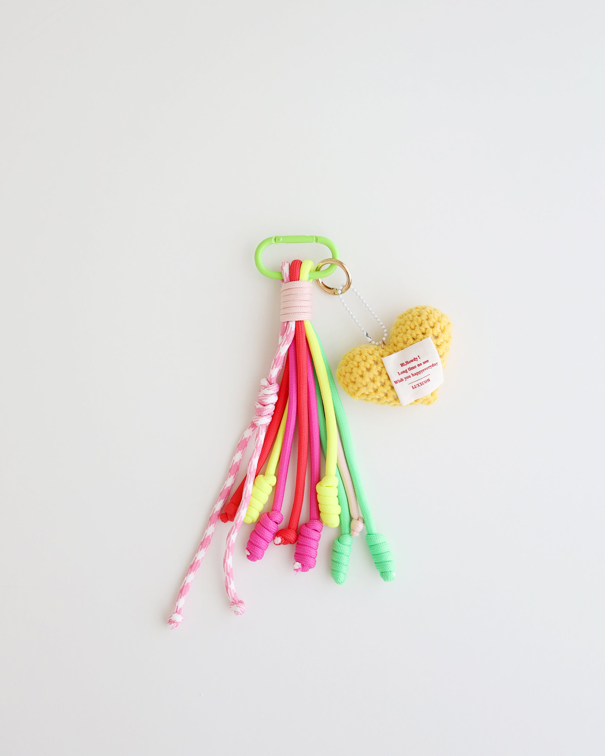 A keychain charm made with multiple knotted cord tassels and a soft yellow crocheted heart which includes a pastel green metal carabiner clip for attaching to bags, keys, or other items. The tassels are made of multi-color cords, and the crochet heart features a stitched label.
