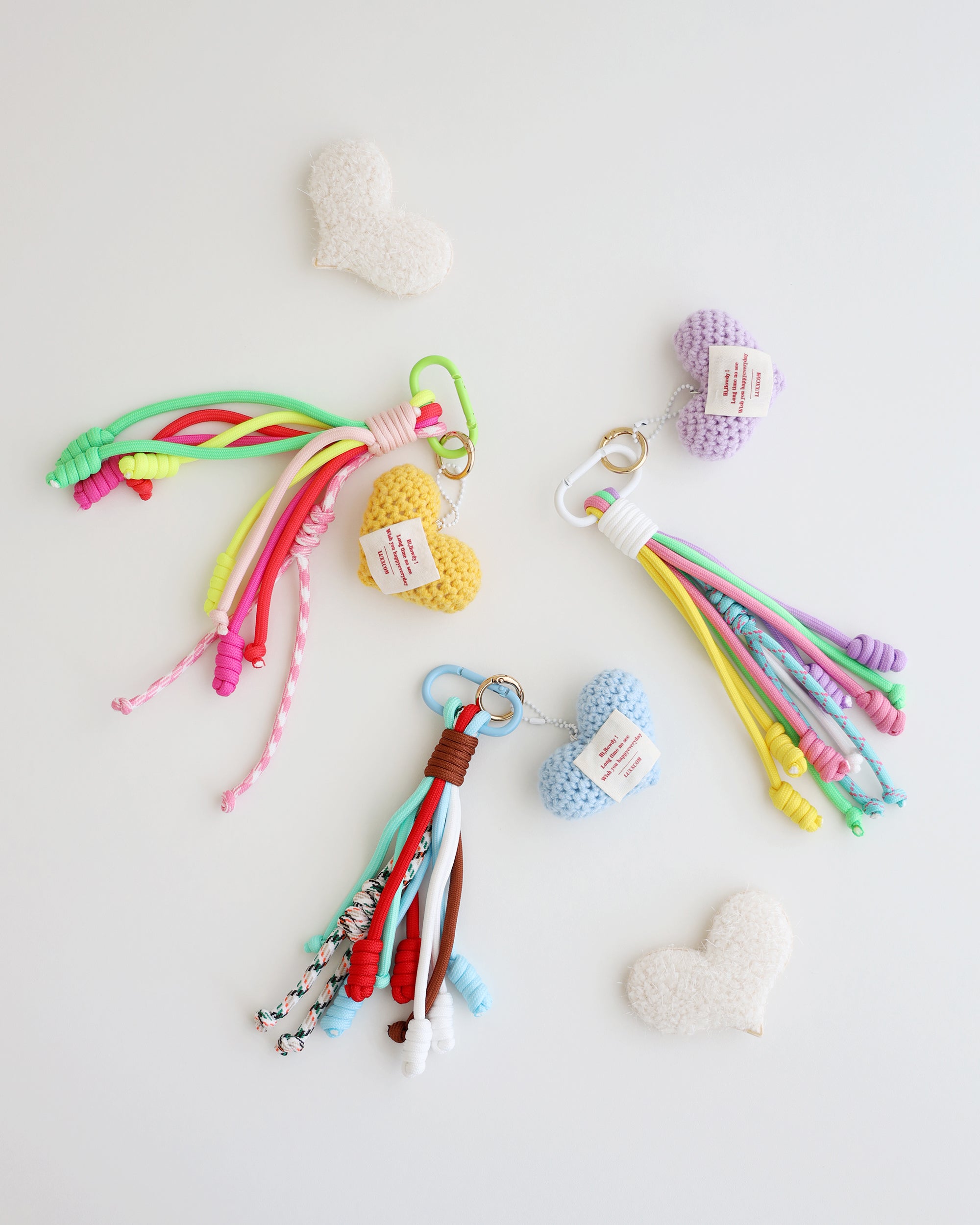 Three keychain charms made with multiple knotted cord tassels and a crocheted heart. Each charm includes a metal carabiner clip for attaching to bags, keys, or other items. The tassels are made of multi-color cords, and the crochet heart features a stitched label displayed in white background with felt heart shaped props.