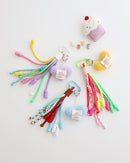 Three keychain charms made with multiple knotted cord tassels and a crocheted heart. Each charm includes a metal carabiner clip for attaching to bags, keys, or other items. The tassels are made of multi-color cords, and the crochet heart features a stitched label displayed in white background with felt cupcake shaped prop.
