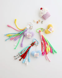 Three keychain charms made with multiple knotted cord tassels and a crocheted heart. Each charm includes a metal carabiner clip for attaching to bags, keys, or other items. The tassels are made of multi-color cords, and the crochet heart features a stitched label displayed in white background with felt cupcake shaped prop.