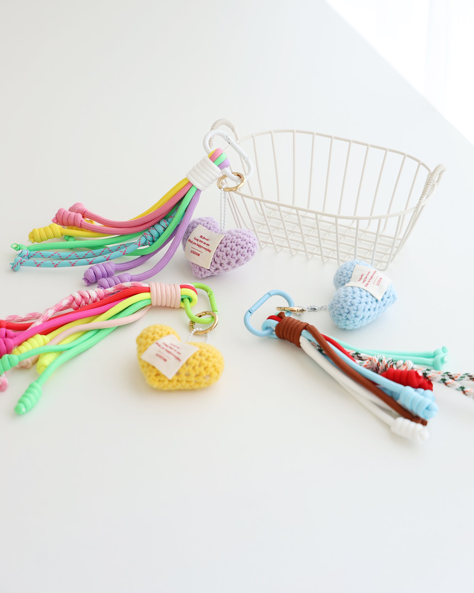 Three keychain charms made with multiple knotted cord tassels and a crocheted heart. Each charm includes a metal carabiner clip for attaching to bags, keys, or other items. The tassels are made of multi-color cords, and the crochet heart features a stitched label displayed in white background with white basket.