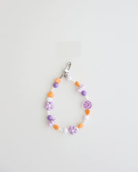 A pastel beaded cell phone strap featuring purple, orange, and white beads with flower and heart accents, plus a small smiley charm. It includes a clear holder insert that slips into a phone case for secure attachment.