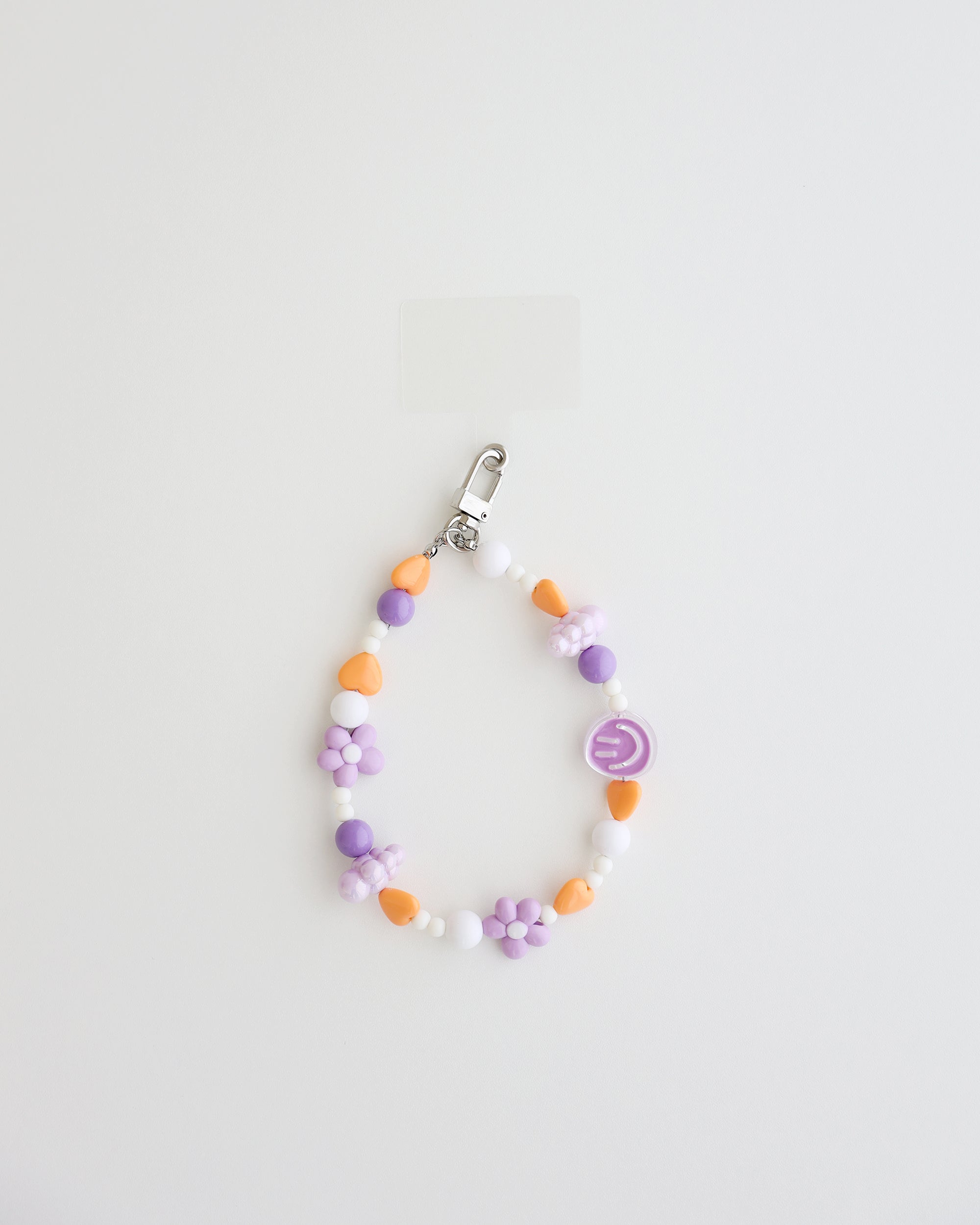 A pastel beaded cell phone strap featuring purple, orange, and white beads with flower and heart accents, plus a small smiley charm. It includes a clear holder insert that slips into a phone case for secure attachment.