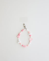A pastel beaded cell phone strap featuring pink, soft pink, and white beads with flower and heart accents, plus a small smiley charm. It includes a clear holder insert that slips into a phone case for secure attachment.