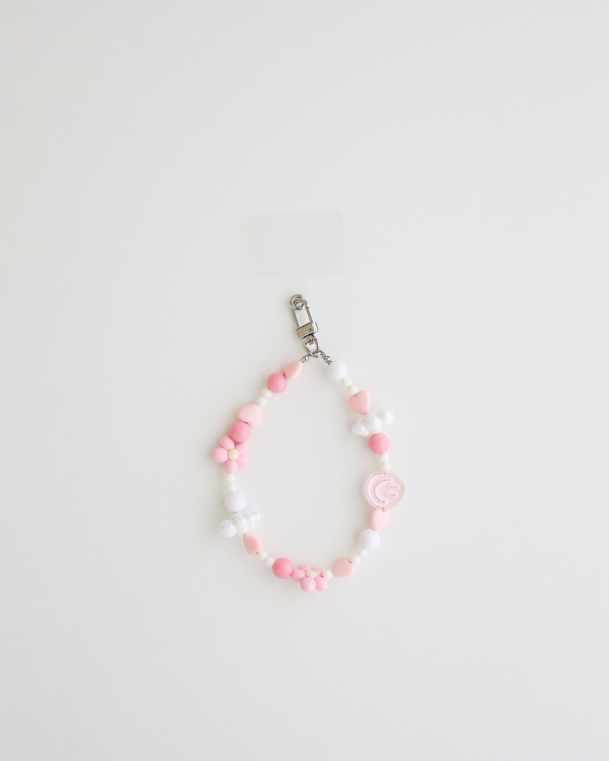 A pastel beaded cell phone strap featuring pink, soft pink, and white beads with flower and heart accents, plus a small smiley charm. It includes a clear holder insert that slips into a phone case for secure attachment.