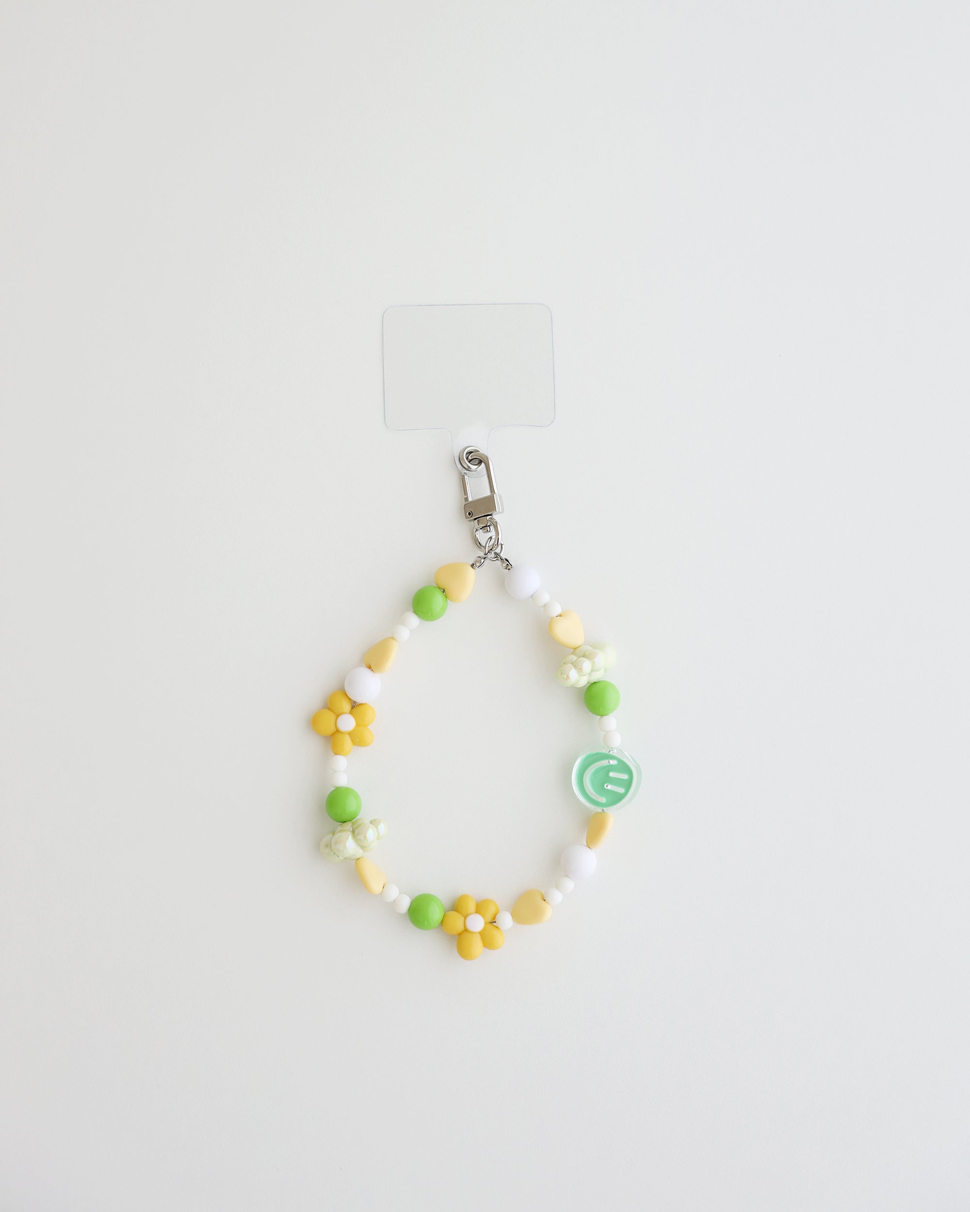 A pastel beaded cell phone strap featuring yellow, soft green, and white beads with flower and heart accents, plus a small smiley charm. It includes a clear holder insert that slips into a phone case for secure attachment.
