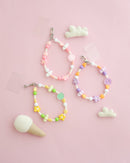 Three colorful beaded bracelets with heart, flower, and smiley face charms in pastel pink, purple, yellow, and green, displayed on a soft pink background with small felt clouds and an ice cream cone prop.