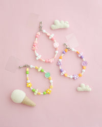 Three colorful beaded bracelets with heart, flower, and smiley face charms in pastel pink, purple, yellow, and green, displayed on a soft pink background with small felt clouds and an ice cream cone prop.