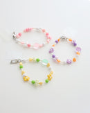 Three colorful cell phone straps made with pastel beads, hearts, flowers, and smiley charms. Each strap has a clear holder designed to slip into a phone case for easy attachment, displayed on a white background.