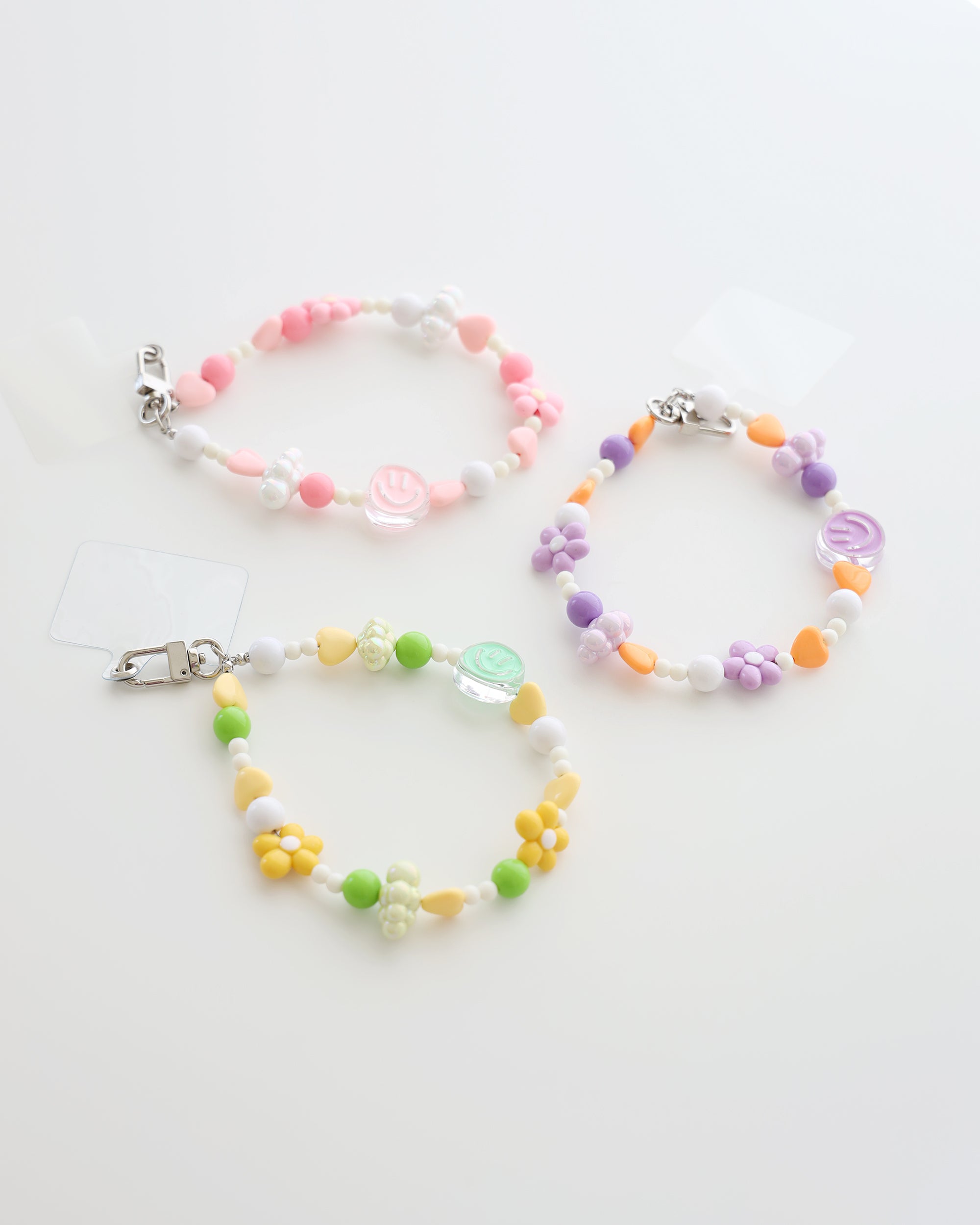 Three colorful cell phone straps made with pastel beads, hearts, flowers, and smiley charms. Each strap has a clear holder designed to slip into a phone case for easy attachment, displayed on a white background.