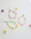 Three colorful cell phone straps made with pastel beads, hearts, flowers, and smiley charms. Each strap has a clear holder designed to slip into a phone case for easy attachment, displayed on a white background with daisy and colorful felt balls.