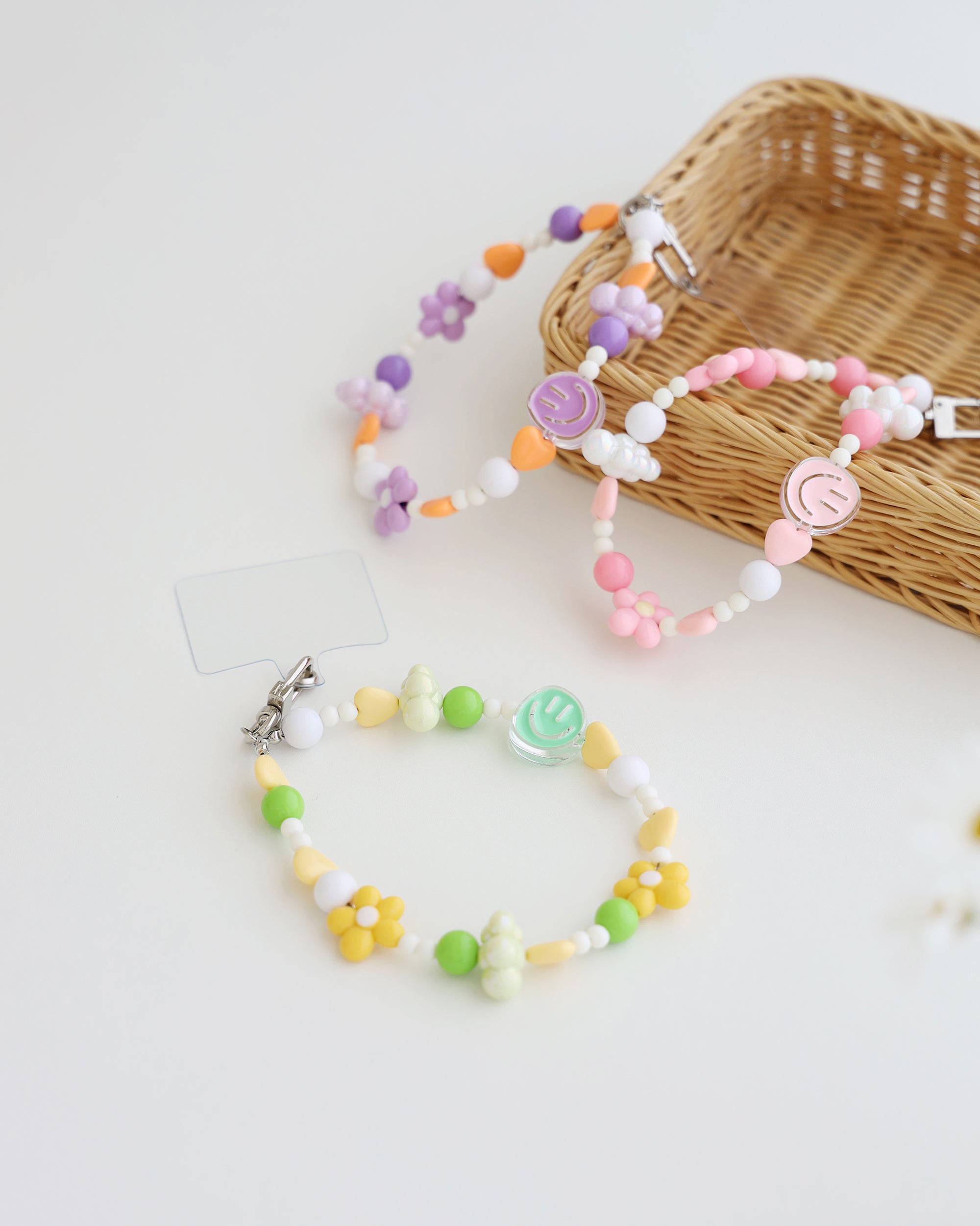 Three colorful cell phone straps made with pastel beads, hearts, flowers, and smiley charms. Each strap has a clear holder designed to slip into a phone case for easy attachment, displayed on a white background with basket.