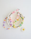 Three colorful cell phone straps made with pastel beads, hearts, flowers, and smiley charms. Each strap has a clear holder designed to slip into a phone case for easy attachment, displayed on a white plate with text around the plate.