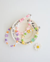 Three colorful cell phone straps made with pastel beads, hearts, flowers, and smiley charms. Each strap has a clear holder designed to slip into a phone case for easy attachment, displayed on a white plate with text around the plate.