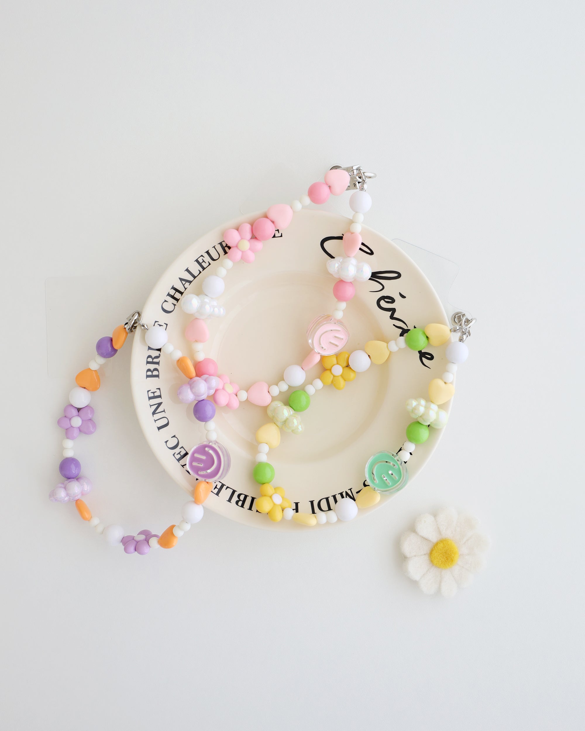 Three colorful cell phone straps made with pastel beads, hearts, flowers, and smiley charms. Each strap has a clear holder designed to slip into a phone case for easy attachment, displayed on a white plate with text around the plate.
