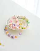 Three colorful cell phone straps made with pastel beads, hearts, flowers, and smiley charms. Each strap has a clear holder designed to slip into a phone case for easy attachment, displayed on a white plate with text around the plate.