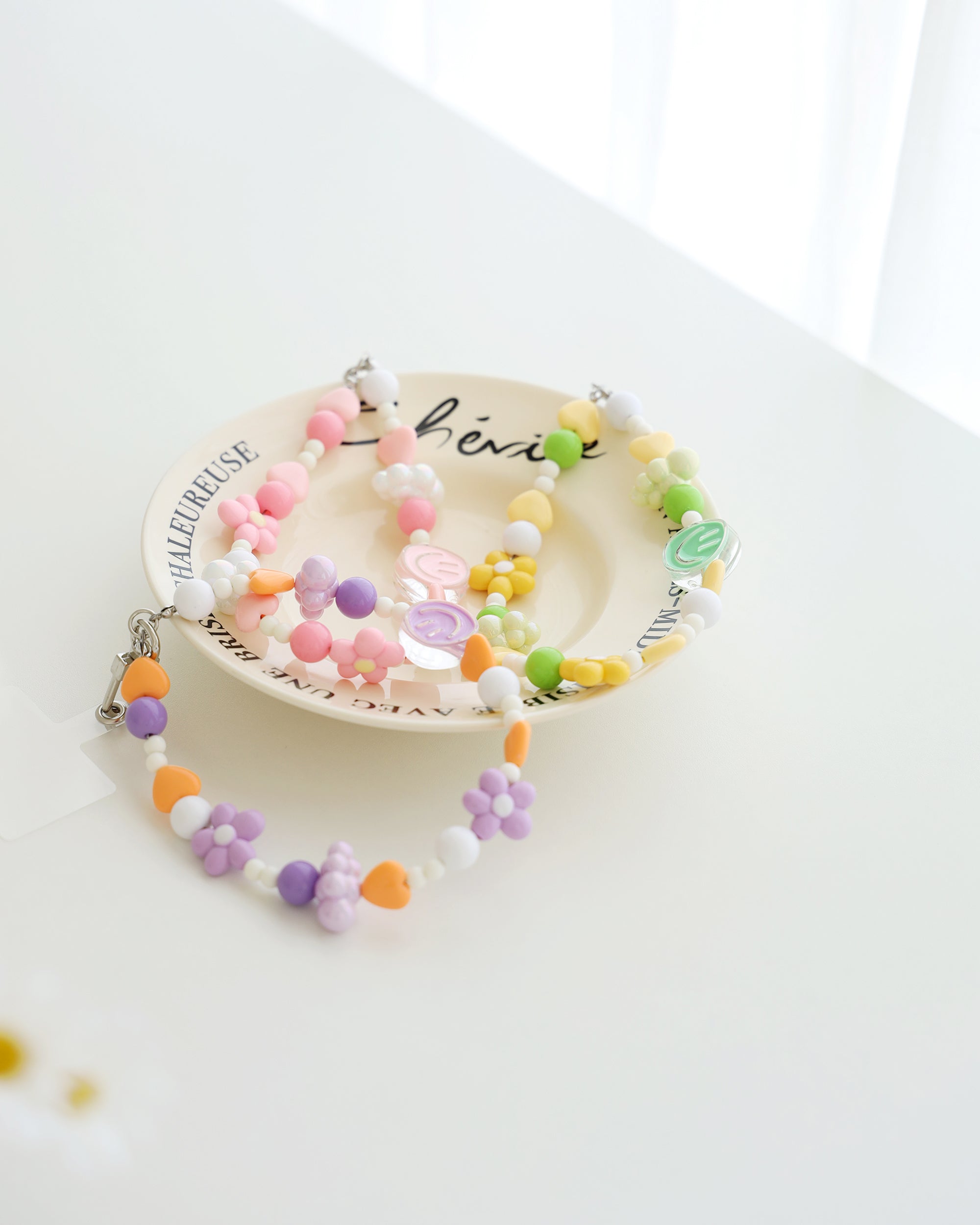 Three colorful cell phone straps made with pastel beads, hearts, flowers, and smiley charms. Each strap has a clear holder designed to slip into a phone case for easy attachment, displayed on a white plate with text around the plate.