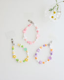 Three colorful cell phone straps made with pastel beads, hearts, flowers, and smiley charms. Each strap has a clear holder designed to slip into a phone case for easy attachment, displayed on white background with daisy flower in a clear vase.