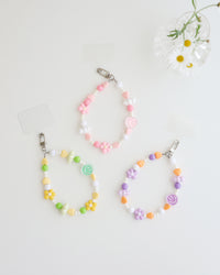 Three colorful cell phone straps made with pastel beads, hearts, flowers, and smiley charms. Each strap has a clear holder designed to slip into a phone case for easy attachment, displayed on white background with daisy flower in a clear vase.