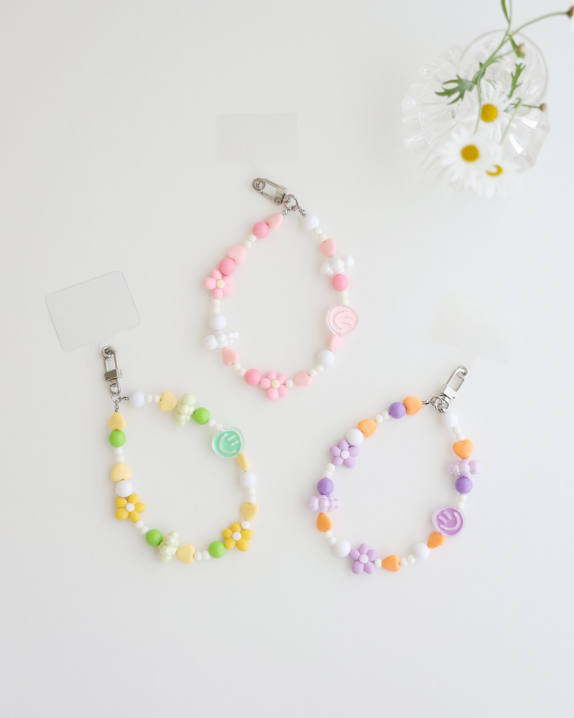 Three colorful cell phone straps made with pastel beads, hearts, flowers, and smiley charms. Each strap has a clear holder designed to slip into a phone case for easy attachment, displayed on white background with daisy flower in a clear vase.