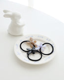 Three black elastic hair ties, each featuring a glossy flower-shaped charm in beige, lavender, and black, displayed on a white plate with handwritten text, next to a small white ceramic bunny.