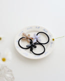 Three black elastic hair ties, each featuring a glossy flower-shaped charm in beige, lavender, and black, displayed on a white plate with handwritten text.
