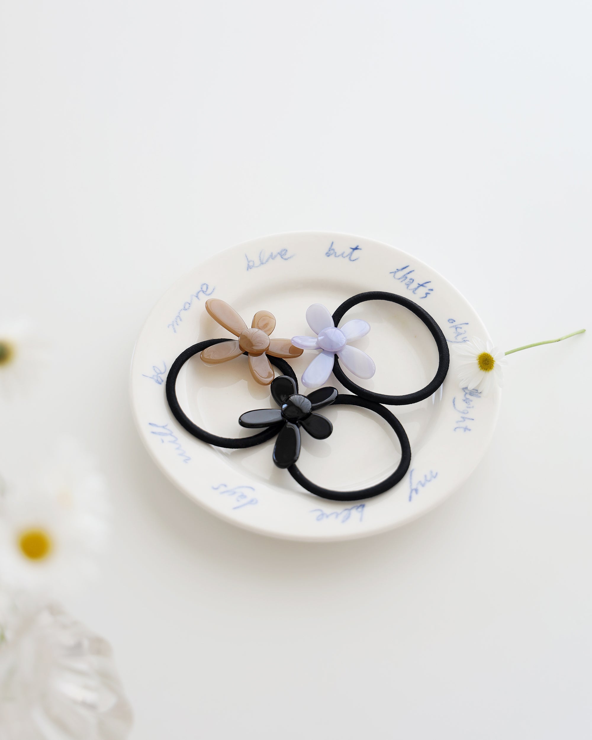 Three black elastic hair ties, each featuring a glossy flower-shaped charm in beige, lavender, and black, displayed on a white plate with handwritten text.