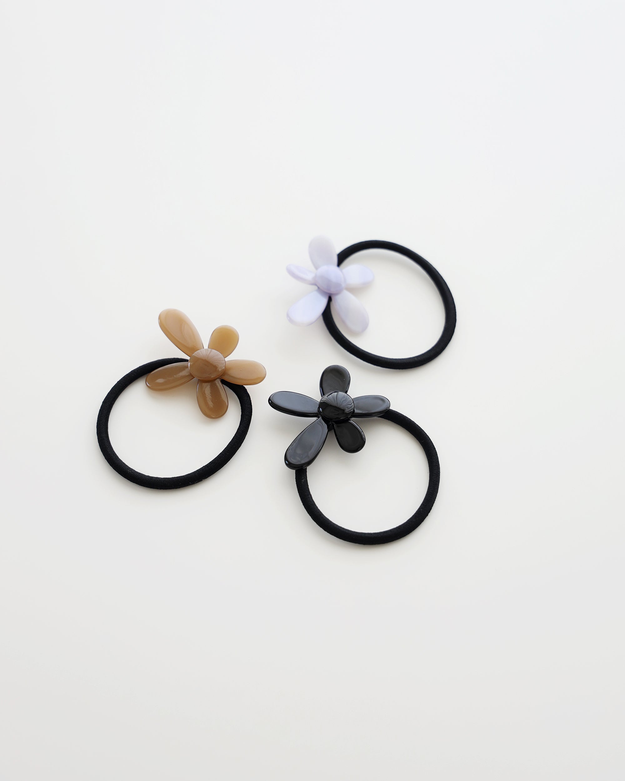Three black elastic hair ties, each featuring a glossy flower-shaped charm in beige, lavender, and black, displayed on a white background.