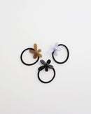 Three black elastic hair ties, each featuring a glossy flower-shaped charm in beige, lavender, and black, displayed on a white background.