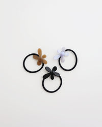 Three black elastic hair ties, each featuring a glossy flower-shaped charm in beige, lavender, and black, displayed on a white background.