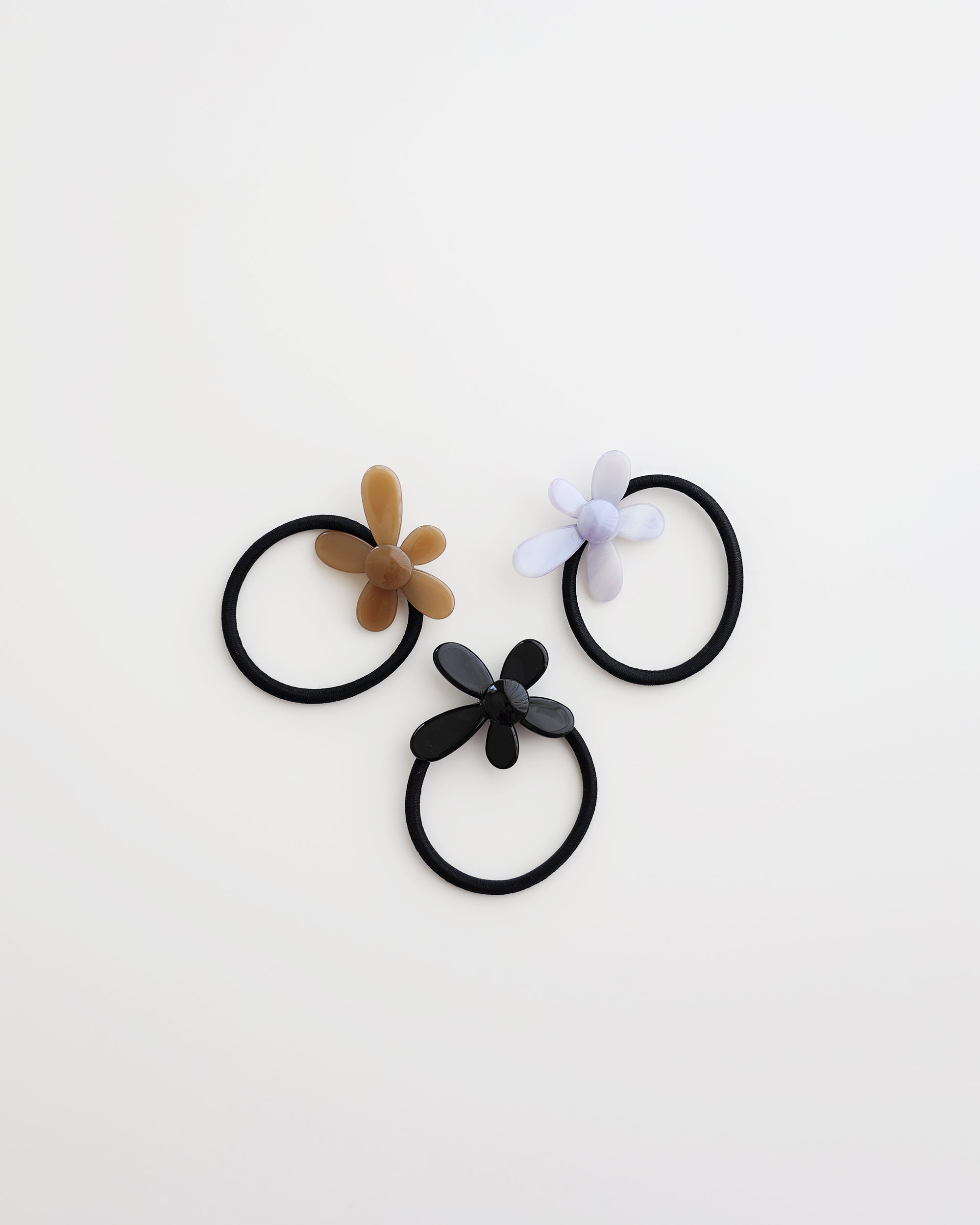 Three black elastic hair ties, each featuring a glossy flower-shaped charm in beige, lavender, and black, displayed on a white background.