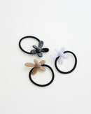 Three black elastic hair ties, each featuring a glossy flower-shaped charm in beige, lavender, and black, displayed on a white background.