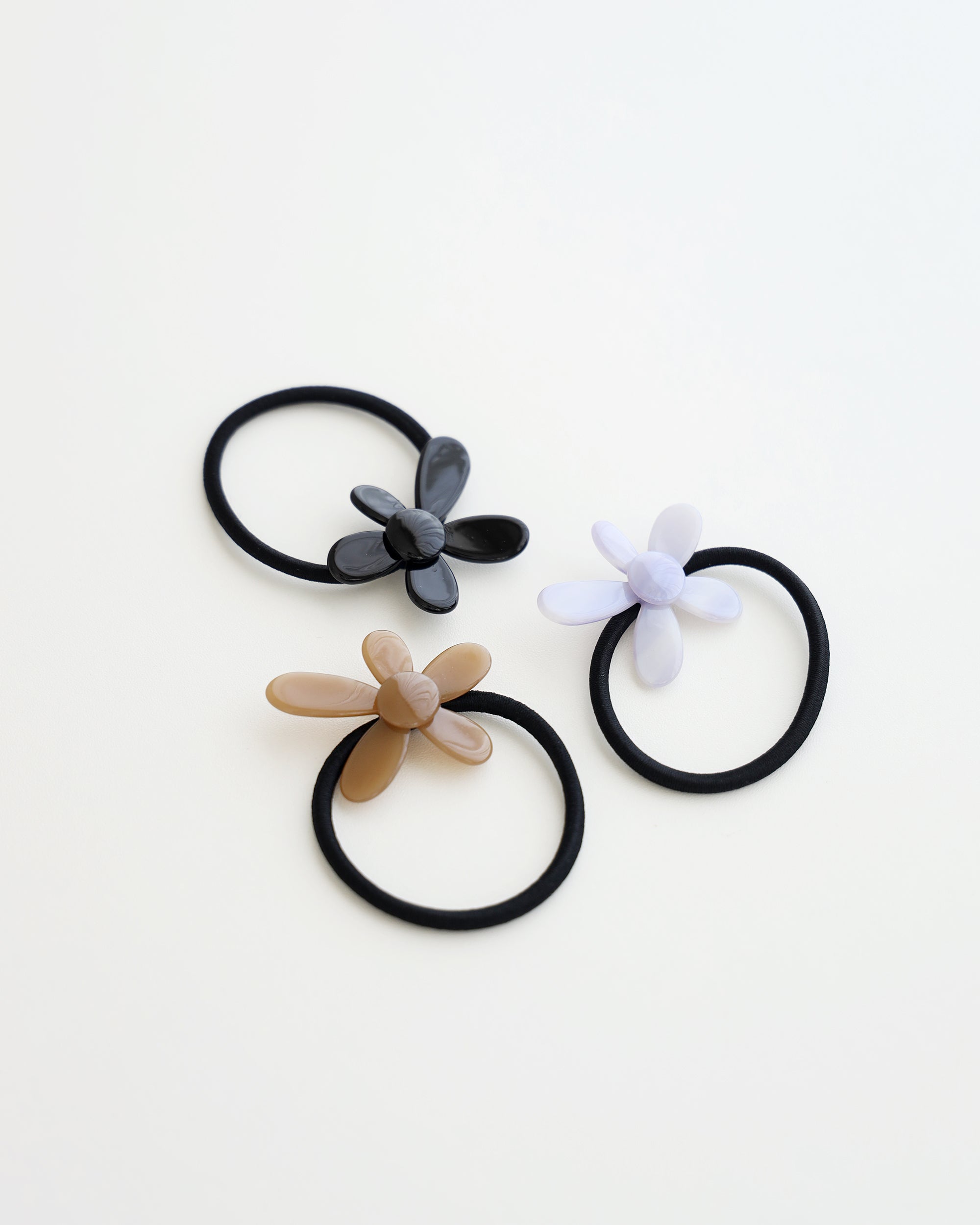 Three black elastic hair ties, each featuring a glossy flower-shaped charm in beige, lavender, and black, displayed on a white background.